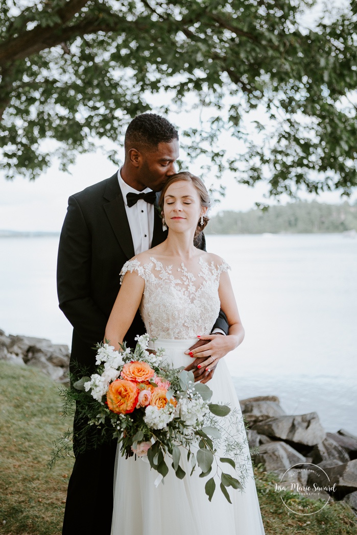 Groom kissing bride on the forehead by the river. African American groom and Caucasian bride. Mariage en Outaouais. Fairmont Le Château Montebello outdoor wedding. Ottawa photographer.