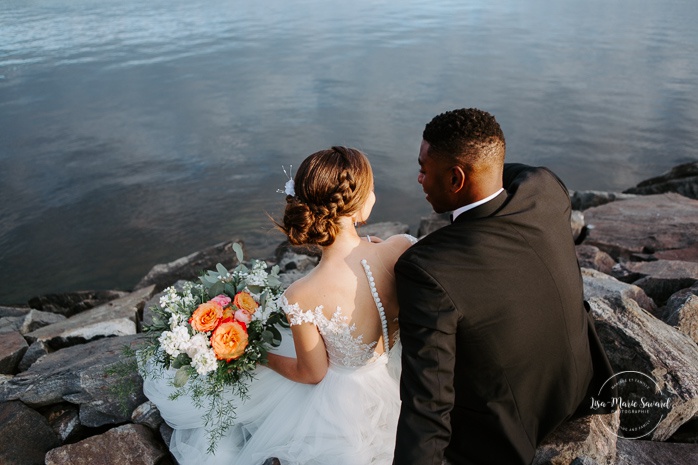 Bride and groom sitting on rocks by the river. African American groom and Caucasian bride. Mariage en Outaouais. Fairmont Le Château Montebello outdoor wedding. Ottawa photographer.