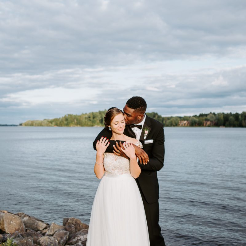 Bride and groom kissing by the river. African American groom and Caucasian bride. Mariage en Outaouais. Fairmont Le Château Montebello outdoor wedding. Ottawa photographer.