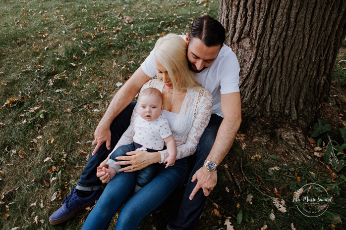 Dad and mom sitting down with baby under a tree. Outdoor family photos with three months old baby boy. Three months milestones. Family photos at the park. Photographe de famille à Montréal.