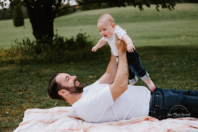 Dad lifting baby in the air on a blanket outside. Outdoor family photos with three months old baby boy. Three months milestones. Family photos at the park. Photographe de famille à Montréal.