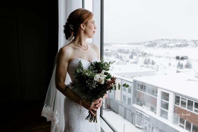 Bride looking at bouquet in front of window. Winter wedding photos. Mariage à Chicoutimi en hiver. Mariage Hôtel Chicoutimi. Photographe de mariage au Saguenay.