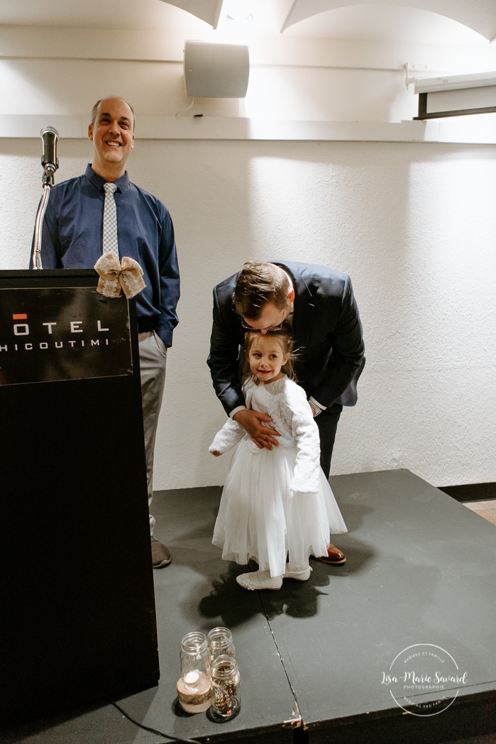 Groom hugging flower girl. Wedding ceremony in dark hall with artificial light. Mariage à Chicoutimi en hiver. Photographe de mariage au Saguenay. Mariage Hôtel Chicoutimi.