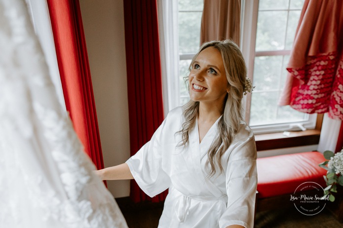 Bride looking at dress in front of window. Bride getting ready with bridesmaids in hotel room. Photographe de mariage en Mauricie. Mariage Le Baluchon Éco-Villégiature. 