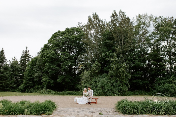 Bride and groom sitting on a bench in front of large trees. Photographe de mariage en Mauricie. Mariage Le Baluchon Éco-Villégiature. 