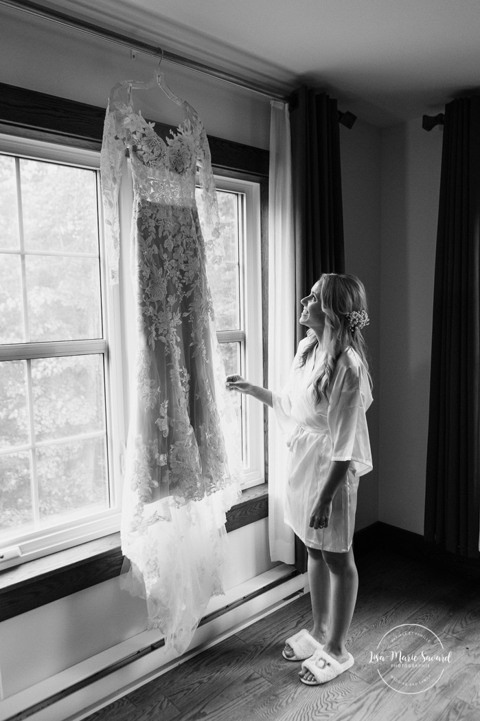 Bride looking at dress in front of window. Bride getting ready with bridesmaids in hotel room. Photographe de mariage en Mauricie. Mariage Le Baluchon Éco-Villégiature. 