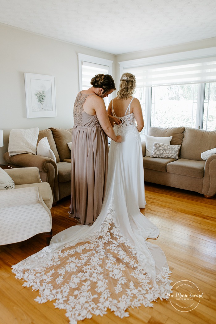 Bridesmaid helping bride button dress. Bride getting with bridesmaids and family at home. Mariage à la Cabane à sucre Constantin. Photographe de mariage à Montréal. Cabane à sucre Constantin wedding. Montreal wedding photographer.