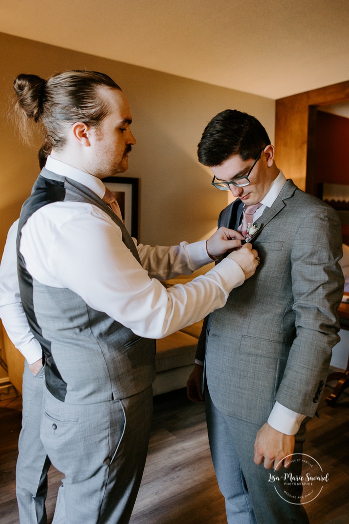 Groom getting ready with two groomsmen in dark hotel room. Photographe de mariage en Estrie. Photographe de mariage Cantons de l'Est. Mariage Estrimont Suites et Spa Orford.