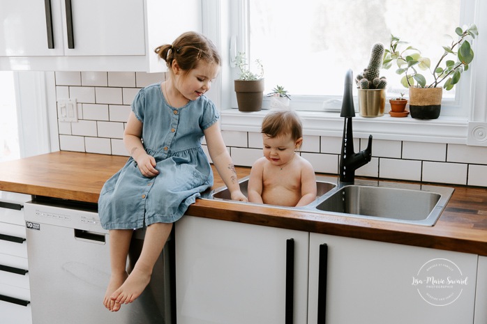 Baby taking bath in kitchen sink with big sister. Baby sink bath with sibling. Kitchen bubble bath. In-home family photos. Lifestyle family photos. Photos de famille à domicile à Montréal. Montreal lifestyle family photos. Photographe famille Montréal. Montreal family photographer.