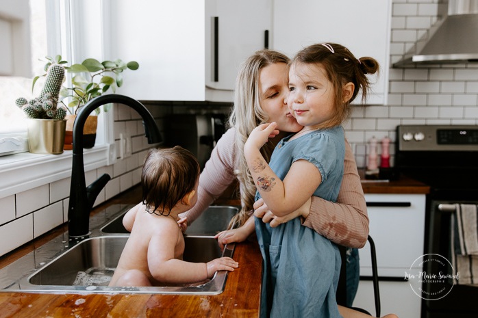 Baby taking bath in kitchen sink with big sister. Baby sink bath with sibling. Kitchen bubble bath. In-home family photos. Lifestyle family photos. Photos de famille à domicile à Montréal. Montreal lifestyle family photos. Photographe famille Montréal. Montreal family photographer.