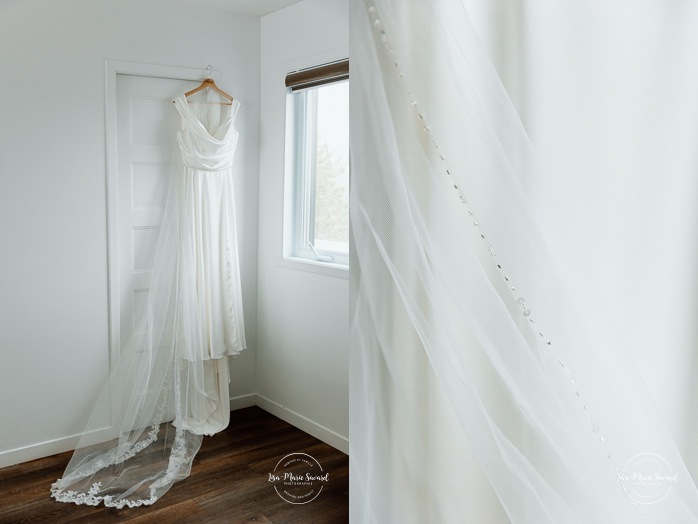 Wedding dress hanging in front of window. Bride getting ready with bridesmaids in hotel room. Mariage à l'Auberge des Îles au Lac-Saint-Jean. Photographe de mariage au Saguenay-Lac-Saint-Jean.