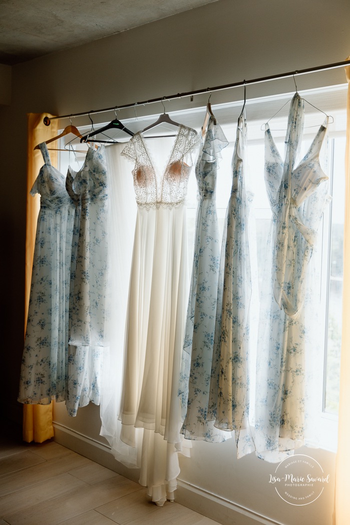 Wedding dresses hanging in front of window. Bride getting ready with bridesmaids. Mariage au bord du Lac-Saint-Jean. Auberge des Îles. Photographe mariage Saguenay-Lac-Saint-Jean